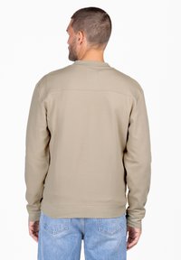 Sudadera beige hecha de una tela suave, con cuello redondo y puños acanalados, y un diseño limpio en la parte posterior. Combinada con jeans de color azul claro.