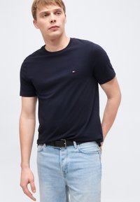 Νεαρός άνδρας που φοράει μπλε σκούρο κοντομάνικο μπλουζάκι και ανοιχτόχρωμα τζιν, στέκεται με το ένα χέρι πίσω από την πλάτη του σε απλό φόντο.