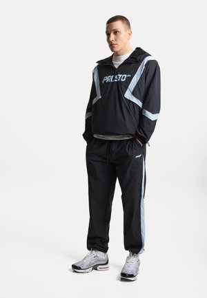 Młody mężczyzna stoi z rękami w kieszeniach, ubrany w czarny i jasnoniebieski dres oraz białe trampki na jednolitym białym tle.