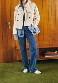 Chaqueta beige de corte recto y doble botonadura con mangas de mezclilla, combinada con jeans azules de pierna recta y zapatillas blancas. El fondo presenta paneles de madera.