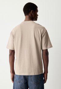 Beige T-shirt met korte mouwen, gemaakt van zacht stof met een relaxte pasvorm. Heeft een ronde hals en een rechte zoom. Draagt met spijkerbroek.