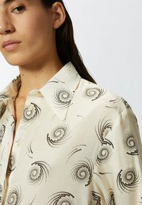 Camicia crema con motivo a spirale nera, design con colletto, chiusura frontale con bottoni e morbida texture del tessuto. Presenta una vestibilità comoda e dettagli sottili.