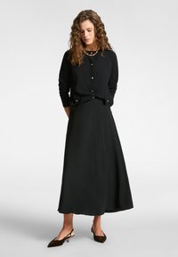 Svart kavaj med knappar tillsammans med en lång svart kjol. Accessoarer inkluderar ett guldhänge och mörka skor med kitten heels.