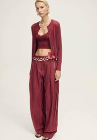 Rotes Schichtoutfit mit einem enganliegenden Tanktop, langem Cardigan und weiten Hosen. Es umfasst einen dekorativen Gürtel mit einer Blumenakzentuierung und runden Elementen.