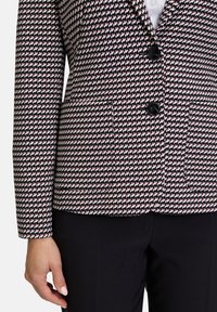 Blazer tissé en noir, blanc et motif pied-de-poule rouge avec un col cranté, deux poches avant et des boutons noirs.