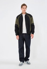 Svart og olivengrønn glidelåsjakke med ståkrage, kombinert med mørkeblå jeans og grå joggesko. Glatt stoff, vanlig passform.