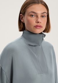 Svetlo šedý pulóver s vysokým golierom, ktorý má zips a textúrovaný panelový detail na prednej strane. Mäkká tkanina s hladkým povrchom.