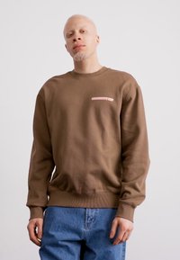 Sudadera marrón con textura acanalada, que presenta un pequeño logo rosa claro en el pecho. Combinada con jeans de mezclilla azules.