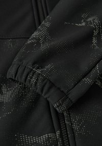 Schwarze Jacke mit elastischem Bündchen, gepunktetem grauen Muster und glatter Stofftextur. Reißverschlussdetail an der Seite sichtbar.