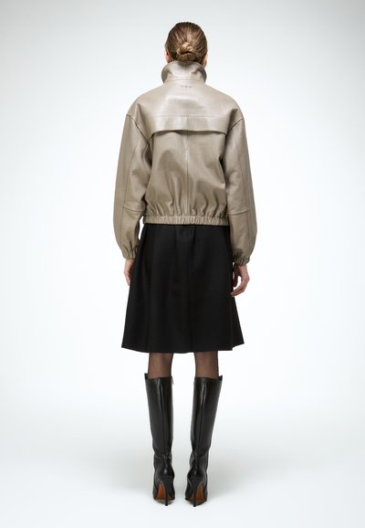 Frau mit Hochsteckfrisur, die eine beige Lederjacke, einen schwarzen knielangen Rock, durchsichtige Strümpfe und schwarze kniehohe Stiefel mit hohen Absätzen trägt, von hinten gesehen.