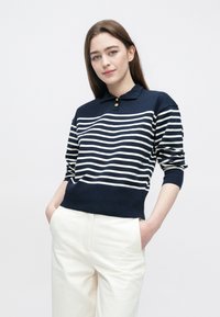 Marineblå og hvit stripet strikket genser med krage, med knappelukking ved halsen og ribbestrikkede mansjetter, kombinert med hvite bukser.