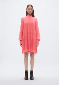 Vestito rosa plissettato con colletto alto e maniche lunghe, caratterizzato da una vestibilità ampia e vita raccolta, abbinato a stivaletti neri.