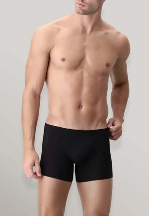 Uomo senza maglietta con fisico tonico che indossa boxer neri, mentre sistema la cintura su sfondo grigio uniforme.