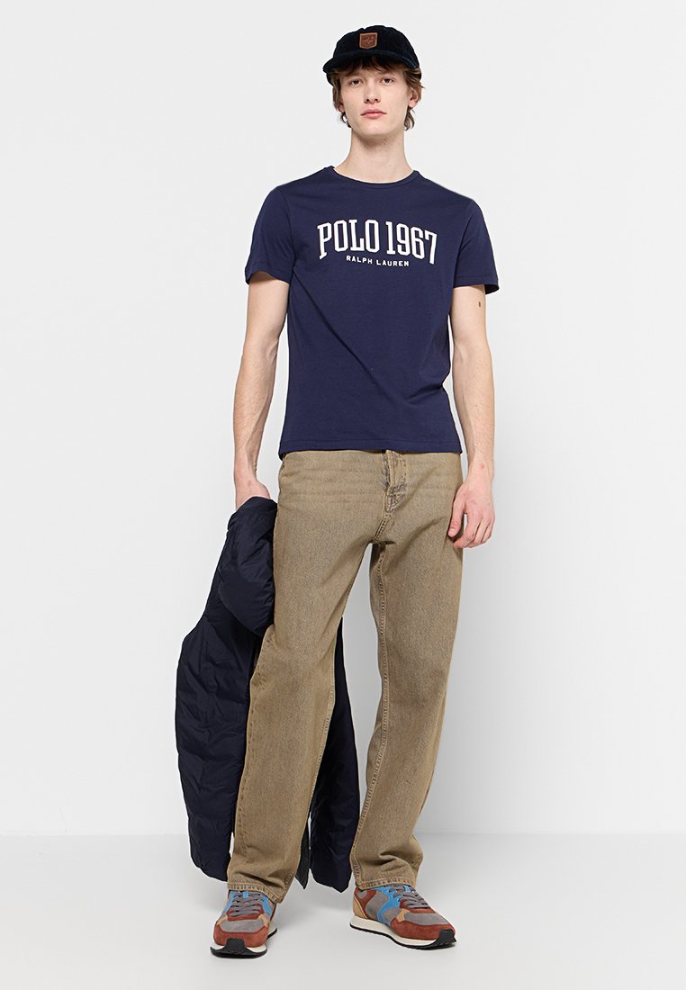 T-shirt in cotone blu navy con testo bianco "POLO 1967 RALPH LAUREN", pantaloni beige, sneakers multicolori e una giacca scura appoggiata al braccio.