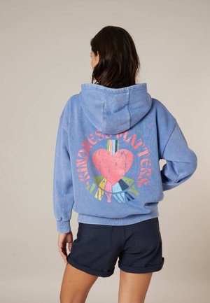 Femme aux cheveux foncés portant un sweat à capuche bleu avec un grand cœur rose et un texte coloré "La gentillesse compte toujours" au dos, associée à un short noir.