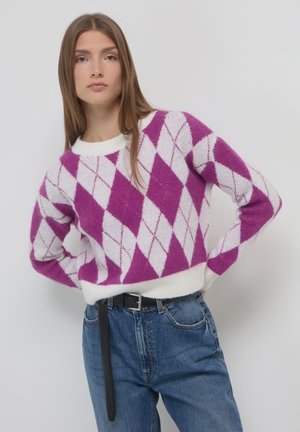 Giovane donna che indossa un maglione argyle viola e bianco e jeans blu, in piedi con le mani sui fianchi contro uno sfondo neutro.