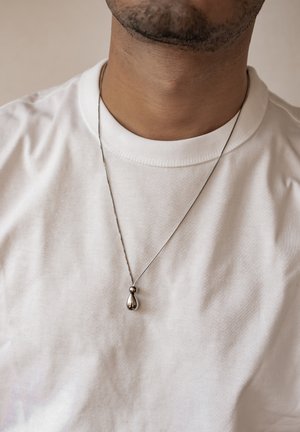 Silver drop pendant on a thin chain, positioned on a white round-neck t-shirt. Smooth texture; subtle shine and minimalistic design.