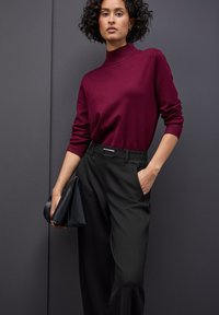 Burgundský pletený svetr s vysokým výstřihem, zkombinovaný s černými kalhotami s vysokým pasem a černou kabelkou na ruku. Hladké materiály, minimalistický design.