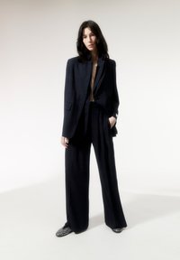 Marineblauw gestreept pak met een op maat gemaakte blazer en wijde broekspijpen. De blazer heeft een gestructureerde kraag en strakke lijnen. Accentschoenen met studs.