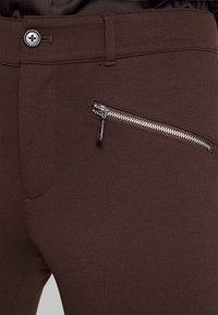 Brązowe dopasowane spodnie o gładkiej fakturze, z boczną kieszenią na zamek i zapięciem na guzik. Zaprojektowane dla ciasnego dopasowania i eleganckiego wyglądu.