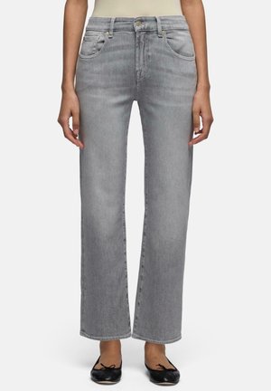 Vrouw draagt lichtgrijze high waist, rechte jeans, gecombineerd met zwarte ballerina's, staand voor een effen achtergrond.