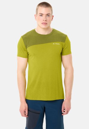 Junger Mann in grünem kurzärmligem Vaude-T-Shirt und marineblauen Shorts, steht vor einem schlichten hellen Hintergrund.