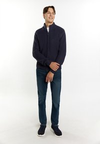 Maglione zip-up a coste di colore navy con colletto alto, abbinato a jeans blu e sneakers scure. Il materiale sembra testurizzato e robusto.