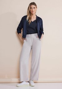 Giacca bomber blu navy, top nero con costina e pantaloni larghi grigi chiari. Sneakers bianche completano l'outfit, posto su uno sfondo neutro.