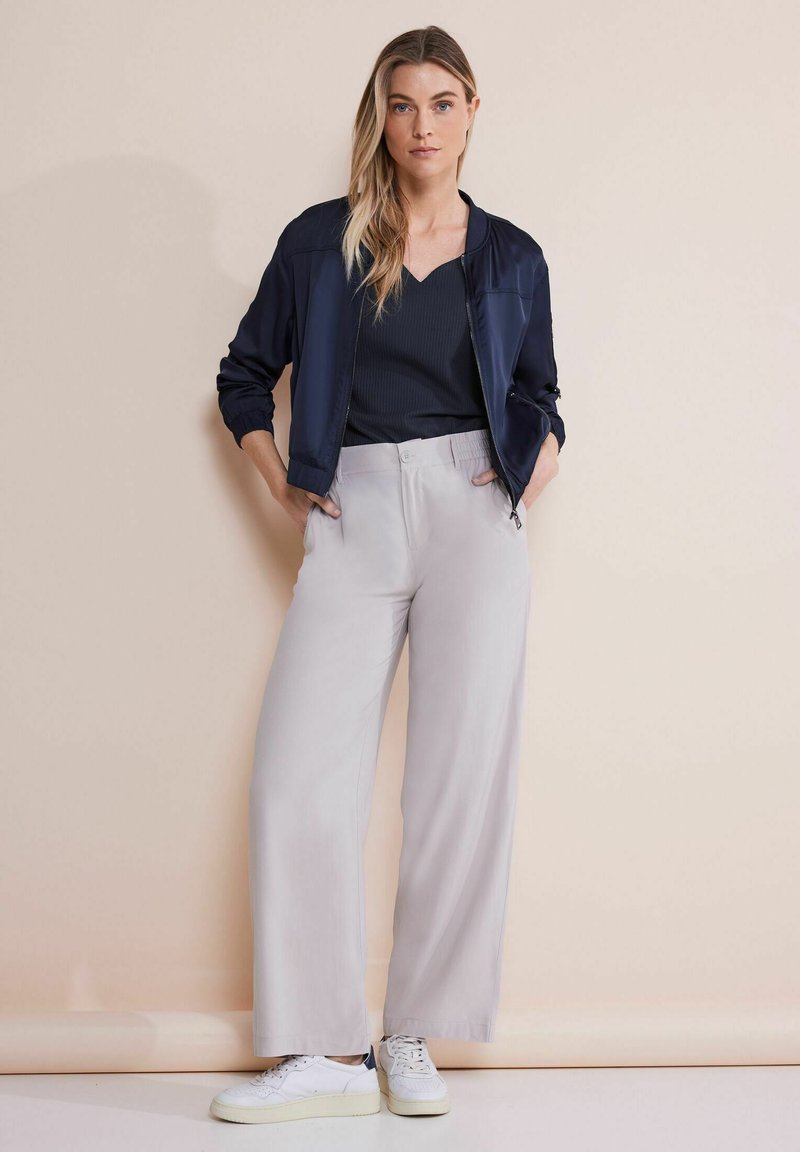 Giacca bomber blu navy, top nero con costina e pantaloni larghi grigi chiari. Sneakers bianche completano l'outfit, posto su uno sfondo neutro.