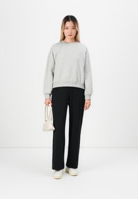 Ung kvinna klädd i en grå sweatshirt, svarta vida byxor, vita sneakers, som håller en liten vit handväska, ståendes mot en slät vit bakgrund.