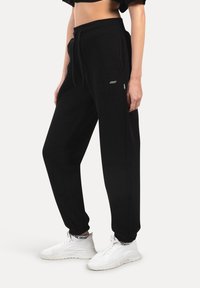 Nainen, jolla on päällään mustat joustavat housut, joissa on narukiristysvyö ja taskut, yhdistettynä valkoisiin lenkkareihin ja mustiin brändättömiin sukkiin.