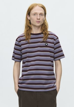 Jeune personne aux longs cheveux roux porte un t-shirt rayé marron et blanc et un pantalon foncé, debout les mains dans les poches devant un fond uni.