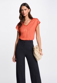 Oranje blouse met korte mouwen en een V-hals met subtiele glinster. Gecombineerd met zwarte wijde broek en een gestructureerde beige clutch.