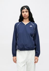 Tengerészkék Nike széldzseki V-nyakkivágással, fehér paszománnyal és rugalmas mandzsettákkal. Könnyű anyagból készült, laza szabású; a modellen világosszürke nadrág van.