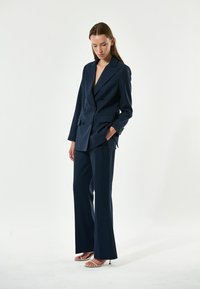Completo blu navy doppiopetto con pantaloni a gamba larga e blazer scollato. Tessuto liscio, design strutturato e dettagli delle tasche frontali ben visibili.