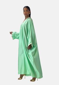Caftan satin vert menthe avec de longues manches, doté d'un décolleté en V et d'une garniture décorative aux poignets. Assorti à des sandales à talons hauts dorées.