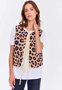 Žena v bezrukávovém béžovém svetru s černými a hnědými leopardími skvrnami přes bílým tričkem a tmavými kalhotami, pózující s rukou u krku.