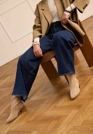 Persoon met een beige blazer, witte top, wijd uitlopende spijkerbroek, beige enkelhoge laarzen en armbanden, zittend op een houten stoel op een parketvloer.