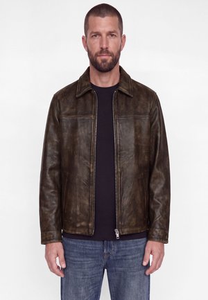Un homme barbu se tient devant un fond blanc. Il porte une veste en cuir marron foncé, une chemise noire et un jean bleu.
