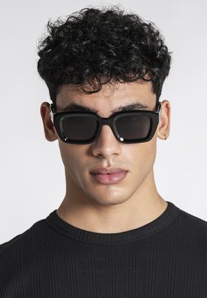 Ung mand med krøllet sort hår iført sort ribstrikket skjorte, rektangulære sorte solbriller og en lille guldring i venstre øre.