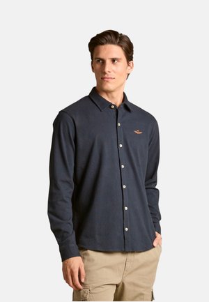 Uomo che indossa una camicia blu navy a maniche lunghe con bottoni e pantaloni cargo beige, in piedi con una mano in tasca, guarda di lato.