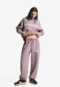 Frau in einem mauvefarbigen Hoodie und Sweatpants mit dem Text "Studio Session 03", die ihren Hoodie anhebt, um ihren Bauch zu zeigen, steht vor einem weißen Hintergrund.