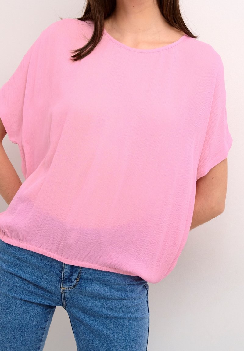 Femme portant un chemisier ample rose clair et un jean bleu, debout les mains derrière le dos sur un fond blanc.