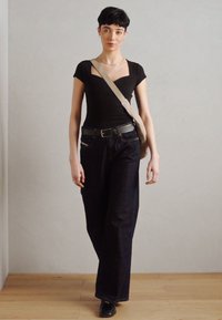 Bodysuit noir à côtes, jean foncé taille haute, ceinture noire et chaussures en cuir. Sac à bandoulière gris. Fond en parquet en bois.