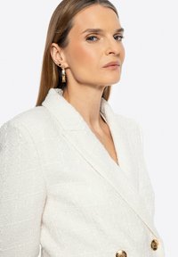 Blazer bianco strutturato con bottoni dorati doppi e colletto a scialle; il modello indossa un orecchino d'oro con un design geometrico.