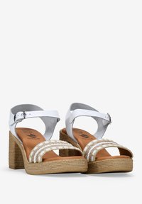 Sandalias blancas de tacón con tiras en el tobillo, que presentan detalles trenzados y una suela de cuña de estilo yute. Acabado de cuero suave con una plantilla marrón.