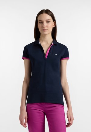 Giovane donna che indossa una polo blu navy con rifiniture rosa e pantaloni magenta, in piedi davanti a uno sfondo bianco uniforme.