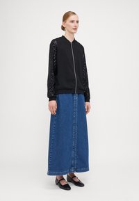Black bomber jacket with sequined sleeves, paired with a long denim skirt, and black slip-on shoes. Clean lines and minimal design.