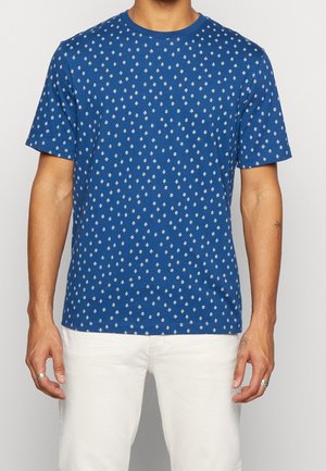 Ciemnoniebieska koszulka z krótkim rękawem, z białym wzorem diamentu, okrągłym dekoltem i miękką bawełnianą tkaniną. Noszona z jasnymi spodniami.