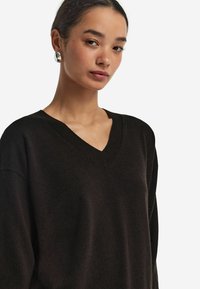 Schwarzer V-Ausschnitt-Pullover aus weichem Material, mit lässiger Passform und geripptem Kragen. Modell trägt silberne Ohrringe, minimaler Hintergrund.